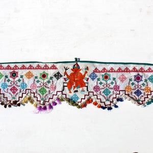 Può includere: Un tendaggio per porta in tessuto bianco con disegni ricamati colorati, tra cui una figura centrale arancione, e un fondo a balze con frange.