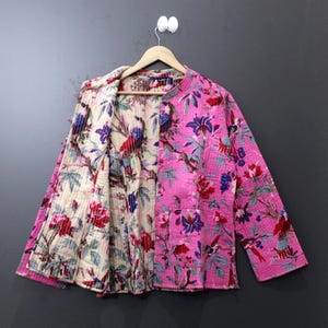 May include: A reversible quilted jacket with a floral pattern. One side is a vibrant pink with red, purple, and blue flowers. The reverse side is a cream color with a similar floral design. The jacket is hanging on a wooden hanger.