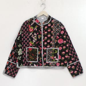 Könnte beinhalten: Eine schwarze gesteppte Jacke mit einem lebendigen Blumenmuster in Pink und Weiß, langen Ärmeln, einem runden Ausschnitt und zwei aufgesetzten Vordertaschen. Die Jacke ist an den Kanten mit einer schwarz-weiß gestreiften Borte versehen.