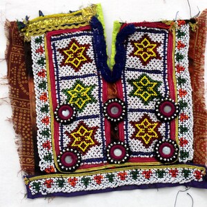 May include: A colorful, embroidered textile fragment with intricate beadwork and mirror accents. The design features geometric star patterns in yellow, green, and white, framed by red and blue borders. The edges are trimmed with gold and purple fabric.