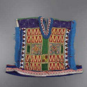 Può includere: Tessuto ricamato e colorato con scollo a V. Il capo presenta un vivace mix di rosso, giallo, verde e blu, con motivi intricati e finiture decorative. Probabilmente è un capo di abbigliamento tradizionale.