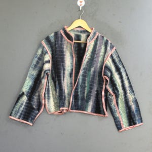 Puede incluir: Una chaqueta corta con un patrón tie-dye en tonos azul oscuro, verde azulado y blanco. La prenda tiene mangas largas, frente abierto y cuello alto, con todos los bordes rematados con un detalle de ribete de tela a rayas rojas y blancas.