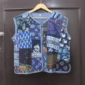 May include: A sleeveless, quilted vest with a patchwork design. The vest features various blue fabric patterns, including floral, geometric, and abstract motifs. It has a V-neck and is lined with a striped fabric. The vest is hanging on a wooden hanger.
