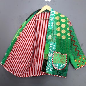 Puede incluir: Chaqueta patchwork con una vibrante mezcla de verde, rojo y blanco. El interior es a rayas rojas y blancas, mientras que el exterior presenta varios parches verdes. La chaqueta está colgada en una percha de madera.