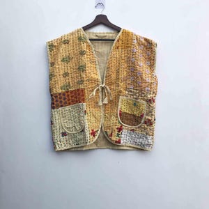 Könnte beinhalten: Eine ärmellose Patchwork-Weste in Gelb-, Beige- und Brauntönen. Die Weste hat verschiedene Stoffmuster, darunter Blumen- und geometrische Designs, und einen Kordelverschluss vorne. Sie hängt an einem Holzbügel vor einer weißen Wand.