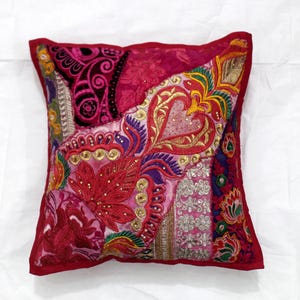 May include: A square, red, pink, and purple patchwork pillow with embroidered floral designs and gold accents.