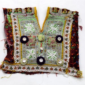 May include: An ornate, vintage textile fragment with a V-neck design. The piece features a central panel of light green fabric with embroidered white floral patterns, framed by silver trim and dark purple circular accents. The edges are adorned with intricate beadwork and a maroon floral patterned fabric.