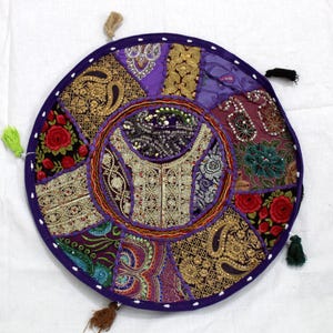 Op de afbeelding: Rond patchwork vloerkussen met een paarse rand en kleurrijke stoffen panelen. Het kussen heeft bloemen- en paisley-patronen in goud, rood, paars en groen. Decoratieve kwastjes zijn aan de rand bevestigd.