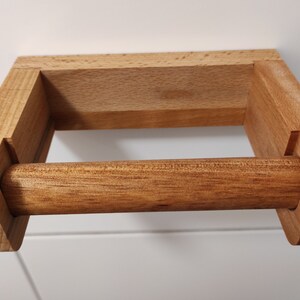 May include: A wall-mounted toilet paper holder crafted from light brown wood. The fixture features a rectangular back plate and two side arms that support a removable horizontal wooden dowel designed to hold a roll of toilet paper.