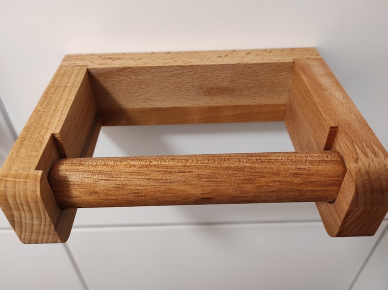 May include: A wall-mounted toilet paper holder crafted from light brown wood. The fixture features a rectangular back plate and two side arms that support a removable horizontal wooden dowel designed to hold a roll of toilet paper.