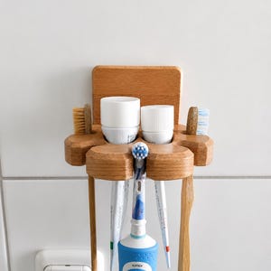 May include: A light brown wooden wall-mounted toothbrush and toothpaste holder featuring a flat back panel, two circular cutouts for toothpaste tubes, and four slots for toothbrushes, including one electric toothbrush.