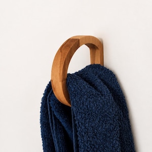 Könnte beinhalten: Ein kreisförmiger Wandhaken aus Holz mit einer glatten, hellbraunen Oberfläche. Der Haken ist an einer flachen Wand befestigt und hält ein gefaltetes, dunkelblaues, strukturiertes Handtuch, das durch die Mitte des Holzrings hängt.