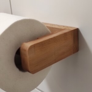 May include: A light brown wooden toilet paper holder mounted to a white tiled wall, holding a standard roll of white toilet paper. The holder features a rectangular arm with rounded edges and a flat base attached to the wall surface.
