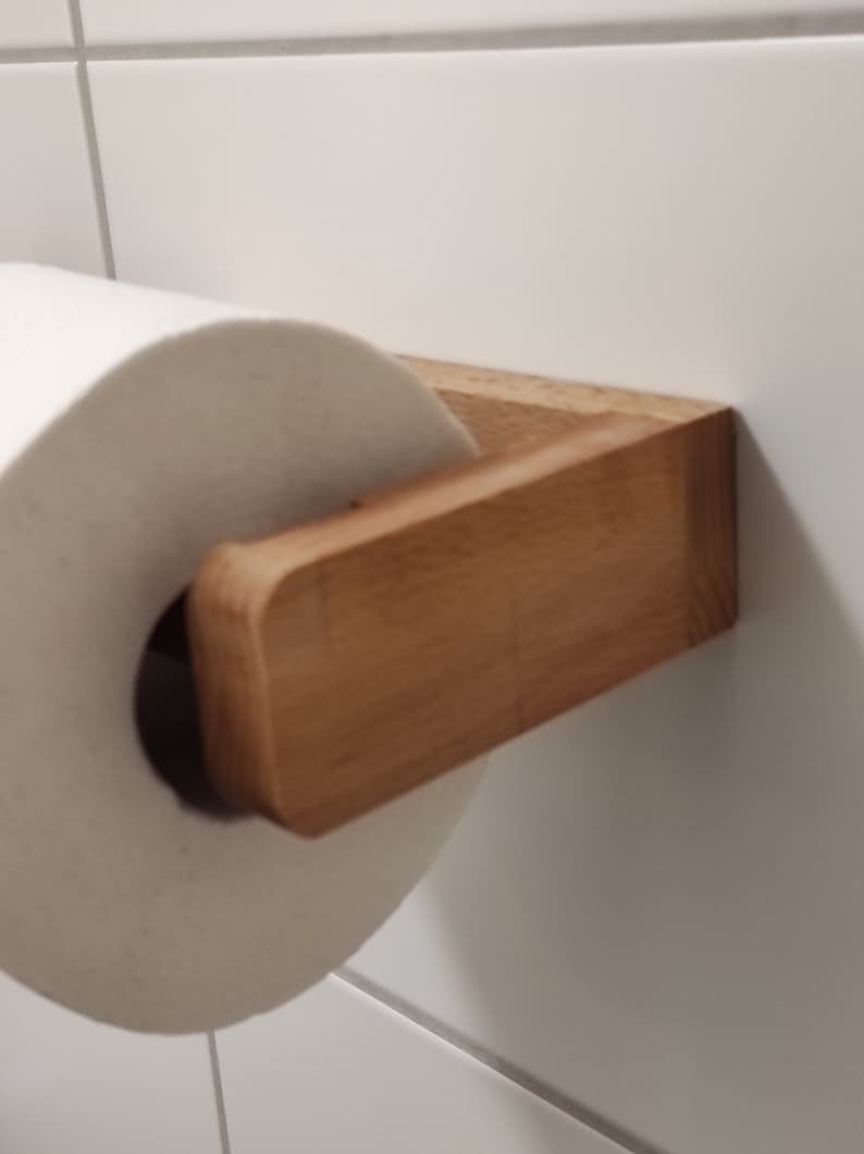 May include: A light brown wooden toilet paper holder mounted to a white tiled wall, holding a standard roll of white toilet paper. The holder features a rectangular arm with rounded edges and a flat base attached to the wall surface.