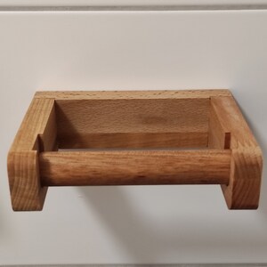 May include: A light brown wooden toilet paper holder featuring a rectangular wall-mounted frame and a horizontal cylindrical rod. The natural wood grain is visible throughout the structure, which is constructed with visible joinery at the corners.