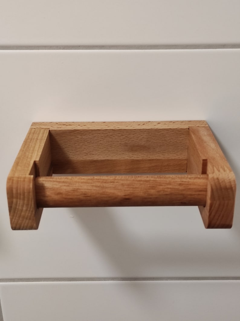 May include: A light brown wooden toilet paper holder featuring a rectangular wall-mounted frame and a horizontal cylindrical rod. The natural wood grain is visible throughout the structure, which is constructed with visible joinery at the corners.