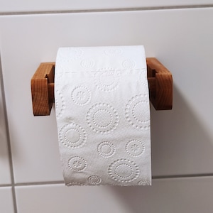 May include: A roll of white toilet paper with an embossed circular pattern hanging on a light brown wooden holder. The paper features repeating designs of small flowers surrounded by dotted rings, suspended on a simple horizontal wooden bracket.