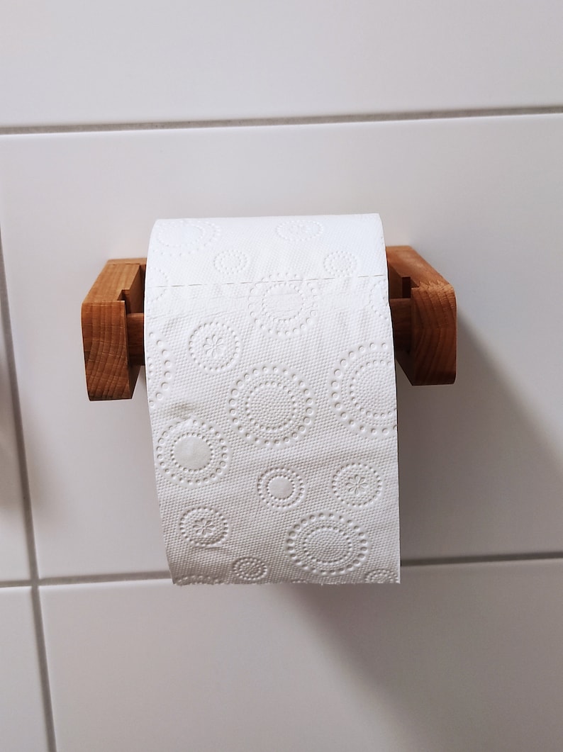 May include: A roll of white toilet paper with an embossed circular pattern hanging on a light brown wooden holder. The paper features repeating designs of small flowers surrounded by dotted rings, suspended on a simple horizontal wooden bracket.