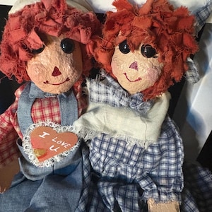 May include: Two handmade rag dolls with red yarn hair, painted faces, and fabric clothing. One doll wears a denim overall and a heart-shaped patch that says "I Love U". The other doll wears a blue and white plaid dress.