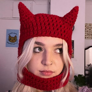 May include: A red crocheted hat with two pointed horns. The hat covers the head and extends down to the neck. The hat is made of a textured yarn and is a vibrant shade of red.
