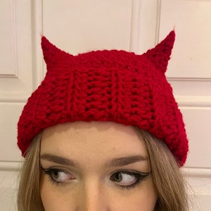 May include: Red knitted beanie with two small horns on top. The beanie is made of a soft, textured yarn.