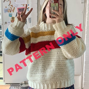 Op de afbeelding: Een witte gehaakte trui met kleurrijke strepen in rood, geel, blauw en wit. De trui heeft een ronde hals en lange mouwen. De tekst "PATTERN ONLY" is over de afbeelding gelegd.
