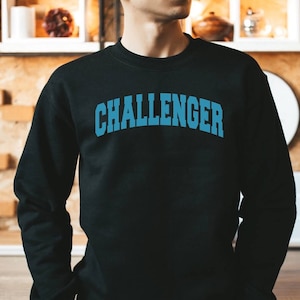May include: Black sweatshirt with the word "CHALLENGER" in blue, arched lettering across the chest. The sweatshirt is long-sleeved and appears to be made of a soft material. The person is wearing the sweatshirt.