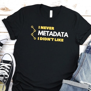 May include: Black t-shirt with a yellow graphic that reads "I never met a metadata I didn't like". The shirt is laid out on a white wooden surface with a pair of black Converse sneakers and a pair of blue jeans.