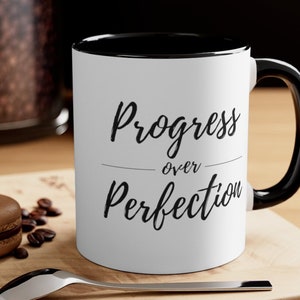 May include: White ceramic mug with a black rim and handle. The mug has black text that reads "Progress over Perfection".