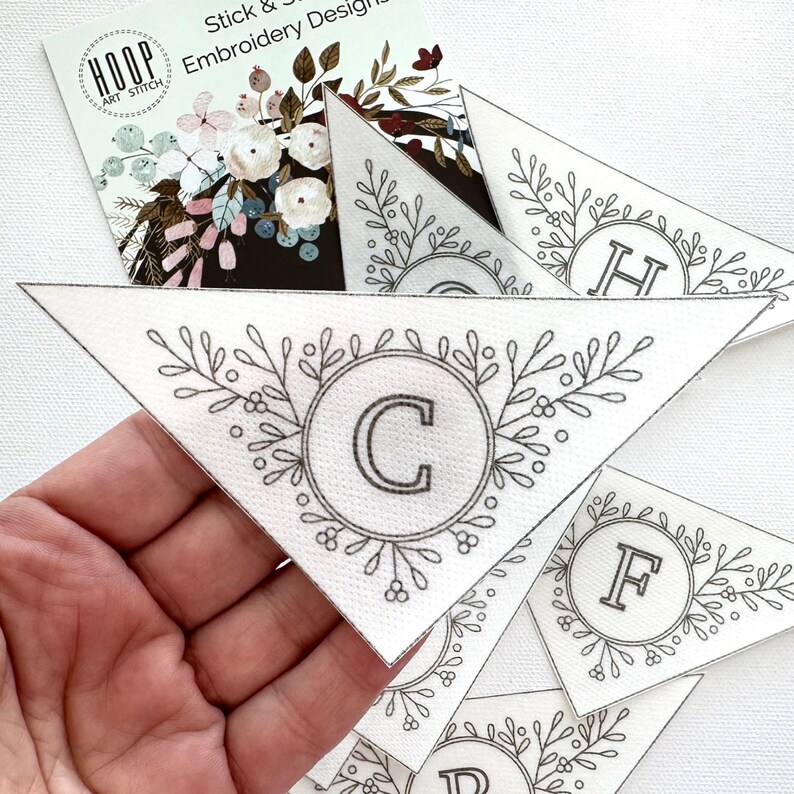 Puede incluir: Dise&ntilde;os de bordado triangulares blancos con una corona floral y una letra en el centro. Los dise&ntilde;os se sostienen en una mano. Una tarjeta de visita con el texto "Hoop Art Stitch Embroidery Designs" est&aacute; en el fondo.