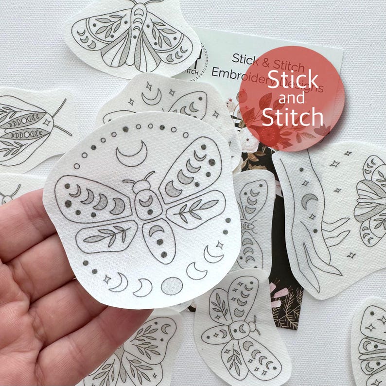 May include: A set of iron-on embroidery designs featuring moths with crescent moons and stars. The designs are printed on white fabric and are ready to be stitched. The text "Stick & Stitch Embroidery Designs" is visible on the packaging.