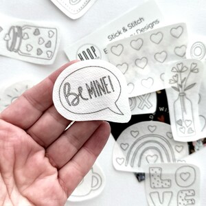 May include: A white iron-on patch with the text "Be Mine!" in a speech bubble. The patch is surrounded by other iron-on patches with various designs, including hearts, flowers, and a rainbow.