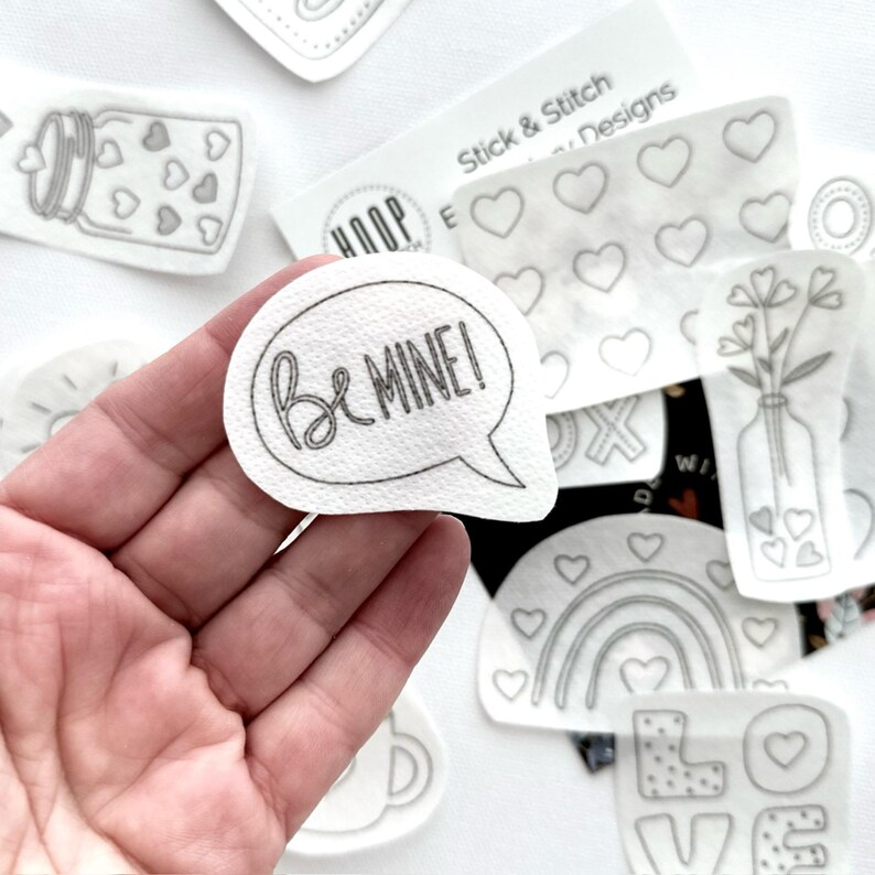 May include: A white iron-on patch with the text "Be Mine!" in a speech bubble. The patch is surrounded by other iron-on patches with various designs, including hearts, flowers, and a rainbow.