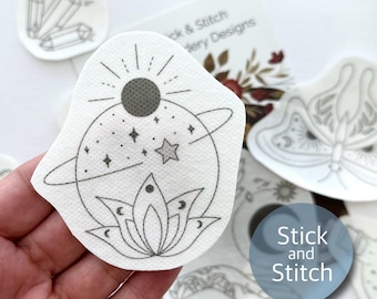 May include: A white iron-on patch with a black line drawing of a planet with a sun, stars, and a lotus flower. The text "Stick and Stitch" is visible in the background.