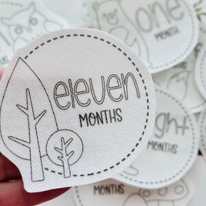 May include: White felt baby milestone circle with black outline and text. The circle features a leaf design with a tree branch and a small tree. The text reads "eleven months".