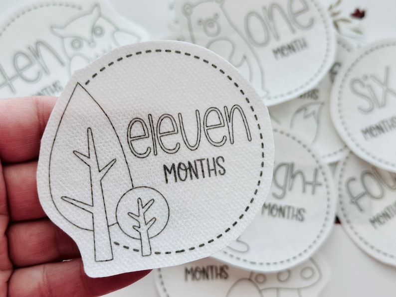 May include: White felt baby milestone circle with black outline and text. The circle features a leaf design with a tree branch and a small tree. The text reads "eleven months".