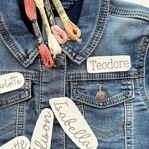 May include: A denim jacket with four spools of embroidery floss in shades of pink, peach, and cream. The jacket has three white fabric patches sewn on the front, each with a name written in black: "Charlotte", "Allison", and "Isabella". A fourth patch is sewn on the collar with the name "Teodore" written in black.