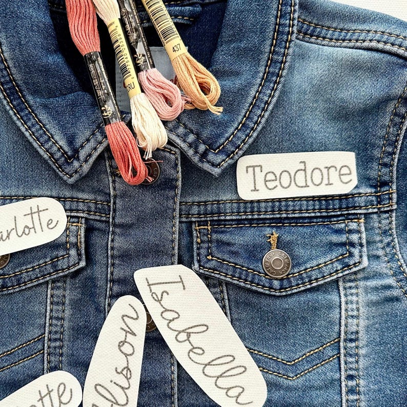 May include: A denim jacket with four spools of embroidery floss in shades of pink, peach, and cream. The jacket has three white fabric patches sewn on the front, each with a name written in black: "Charlotte", "Allison", and "Isabella". A fourth patch is sewn on the collar with the name "Teodore" written in black.