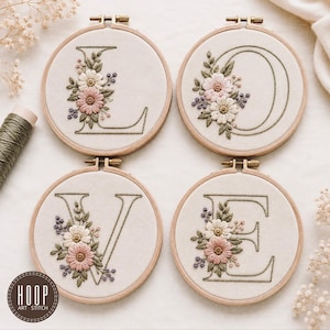 May include: Four circular wooden embroidery hoops containing off-white fabric. Each hoop features a hand-stitched letter spelling LOVE in dark green thread, accented with embroidered pink and white flowers, green leaves, and small purple buds.