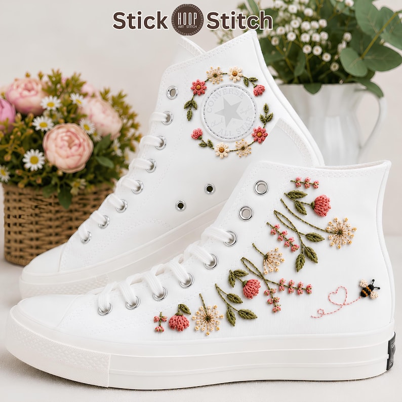 Puede incluir: Un par de zapatillas altas blancas con intrincados bordados florales. El zapato superior muestra una corona circular de flores alrededor del logo, mientras que el zapato inferior presenta una enredadera de flores rosas y blancas.