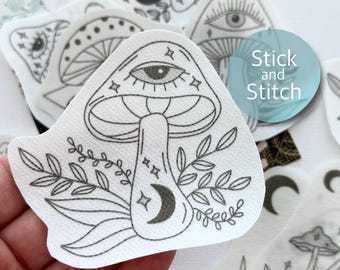 May include: A black and white line drawing of a mushroom with an eye and a crescent moon on the stem. The mushroom is surrounded by leaves and the words "Stick and Stitch" are in the background.