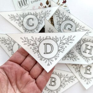 Puede incluir: Piezas triangulares de tela con letras bordadas y dise&ntilde;os de coronas florales. La letra "D" se muestra de forma destacada en una pieza, sostenida por una mano. Otras letras, incluyendo "C", "G", "H", "E" y "B", son visibles en las otras piezas.
