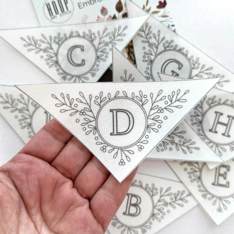 Puede incluir: Piezas triangulares de tela con letras bordadas y dise&ntilde;os de coronas florales. La letra "D" se muestra de forma destacada en una pieza, sostenida por una mano. Otras letras, incluyendo "C", "G", "H", "E" y "B", son visibles en las otras piezas.