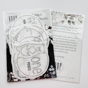 May include: A set of 10 white felt patches with black embroidery designs for baby's first year milestones. The patches feature designs like a bird, a bear, and a cross. The text on the patches reads "One Month".