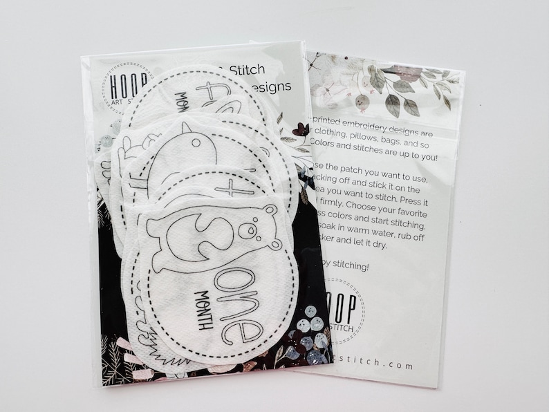 May include: A set of 10 white felt patches with black embroidery designs for baby's first year milestones. The patches feature designs like a bird, a bear, and a cross. The text on the patches reads "One Month".