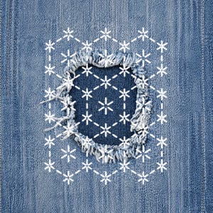 May include: Close-up of a denim patch with a distressed edge and a white embroidered snowflake pattern. The patch is sewn onto a blue denim fabric, creating a contrast in texture and color. The design features a geometric arrangement of snowflakes.