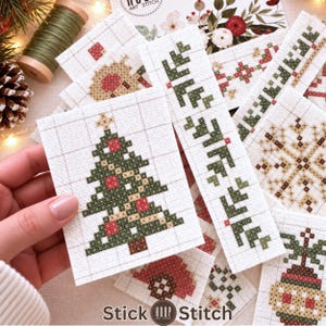 Può includere: Una collezione di pezzi di tessuto bianco con vari disegni natalizi a punto croce, tra cui un albero di Natale verde con ornamenti rossi, un fiocco di neve e bordi decorativi, il tutto con il testo Stick Hoop Stitch.