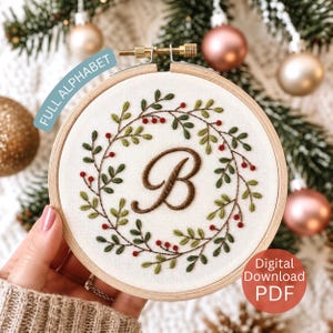 May include: A circular wooden embroidery hoop containing white fabric with a hand-stitched wreath of green leaves and small red berries surrounding a large, brown, cursive letter B. The text FULL ALPHABET and Digital Download PDF are visible.