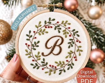 May include: A circular wooden embroidery hoop containing white fabric with a hand-stitched wreath of green leaves and small red berries surrounding a large, brown, cursive letter B. The text FULL ALPHABET and Digital Download PDF are visible.