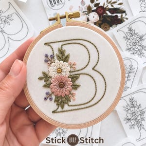 May include: A circular wooden embroidery hoop containing white fabric with a hand-stitched letter B in olive green thread. The letter is decorated with embroidered flowers in shades of white, dusty pink, and purple, surrounded by green leaves.